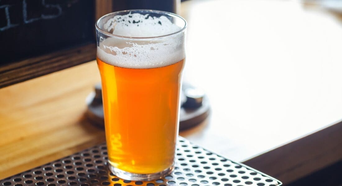 A glass of amber beer with a frothy head sits on a metal mat on a wooden countertop. mosaic hops brew recipe