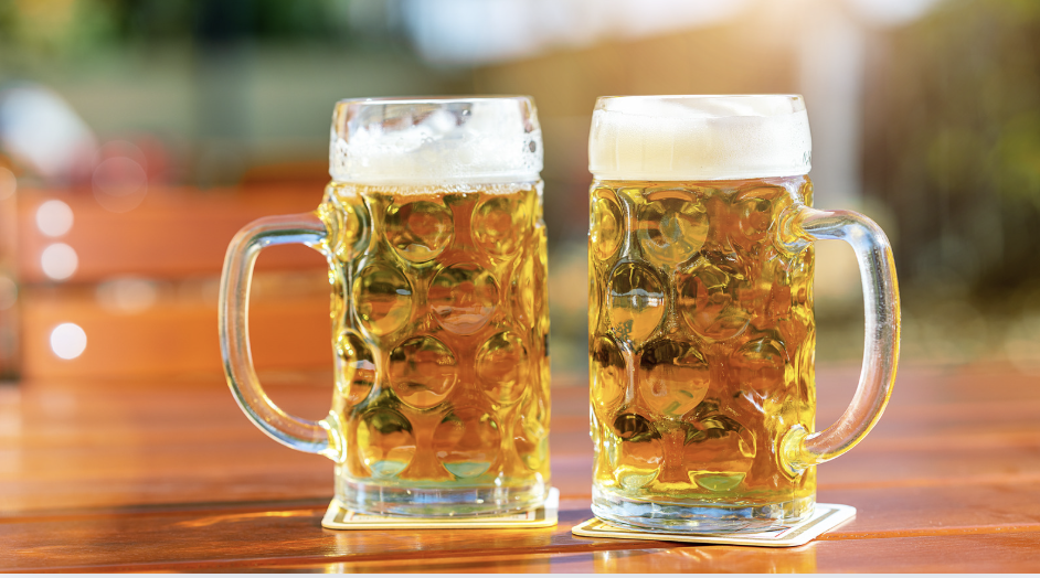 Two large beer mugs filled with light beer on a wooden table, placed on coasters, with a blurred outdoor background and sunlight.